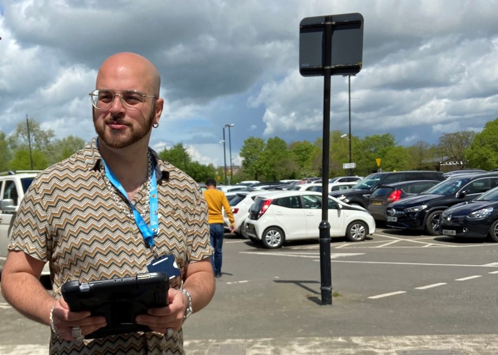 A man stands at the edge of a car park holding a tablet device. He is wearing glasses, looking past the camera and a breeze is blowing a light blue lanyard that he is wearing.