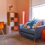 A bright, clean and tidy corner of a child's bedroom. There is a blue sofa, a shelving unit, a desk and chair and a large window.