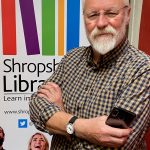 A man with a white beard is smiling at the camera, with arms folded and is holding a mobile phone in one hand. He is wearing glasses and a checked shirt. There is a colourful illustration of books behind him on a poster.