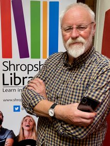 A man with a white beard is smiling at the camera, with arms folded and is holding a mobile phone in one hand. He is wearing glasses and a checked shirt. There is a colourful illustration of books behind him on a poster.