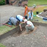 An image of the Shrewsbury Castle dig team discussing layers in part of the trench, trying to understand the relationships between different elements of the archaeology.