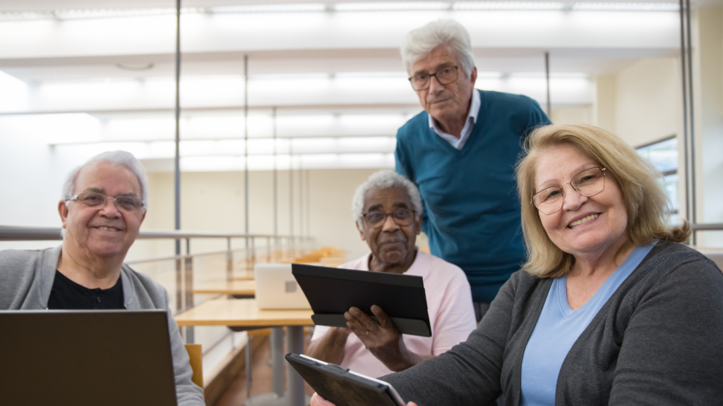 Three people are sitting down and holding tablet devices whilst another person stands nearby and looks on. Everybody is smiling. They are indoors, in a large, white building with a row of empty desks in the background.