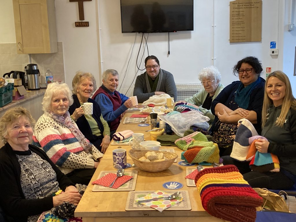 A group of people sit around a table smiling. There is knitting in front of them.
