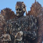 A statue of a winged angel made from knives with blue sky and shops in the background.