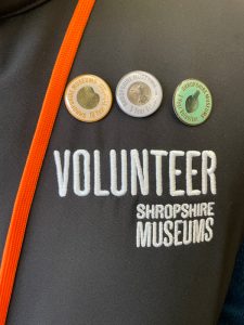 Three metal coloured badges pinned to clothing with the words Volunteer Shropshire Museums