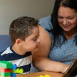 A woman is showing something to a young boy on a screen, there are some toys in the foreground.