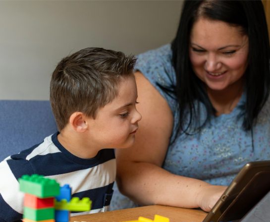 A woman is showing something to a young boy on a screen, there are some toys in the foreground.