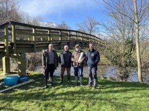 four people standing on grass with a river and a bridge behind