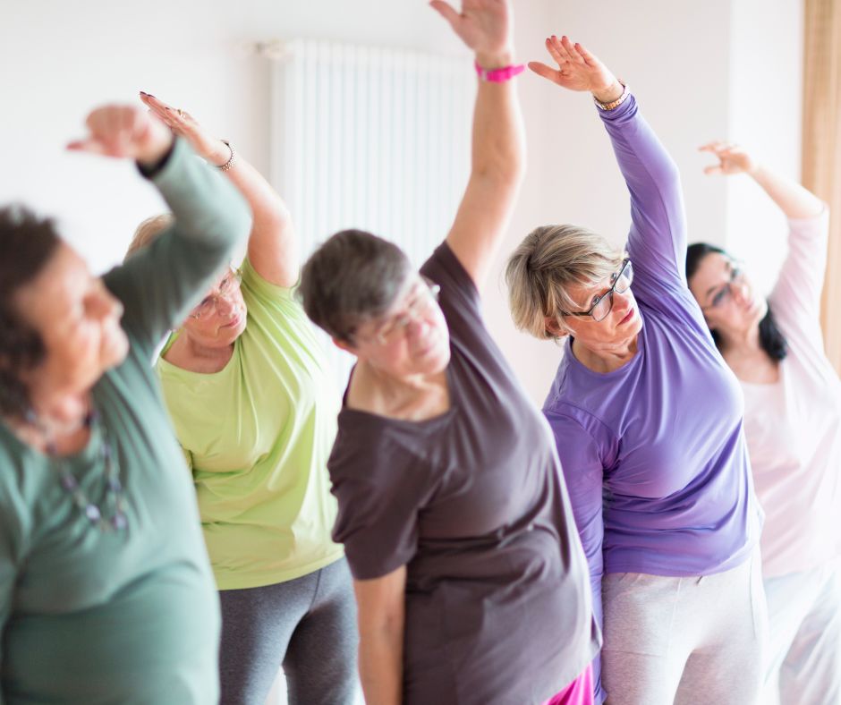 A group of white females are in a brightly lit room, facing in one direction and doing light exercise stretches
