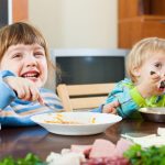 Two young white children sit at a table with a bowl of food each. They are smiling and looking past the camera