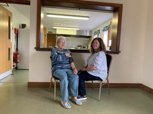 Two people are sitting on chairs in a village hall. One is having a blood pressure check taken, holding the device to her arm. The other person is holding the end of the machine. Both are smiling. 