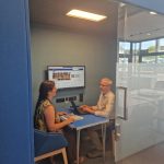 A man and a woman sitting at a desk inside the sound-proof pod with a computer screen in use in the background