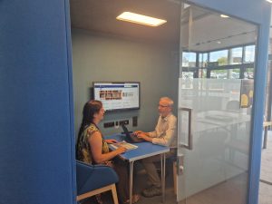 A man and a woman sitting at a desk inside the sound-proof pod with a computer screen in use in the background