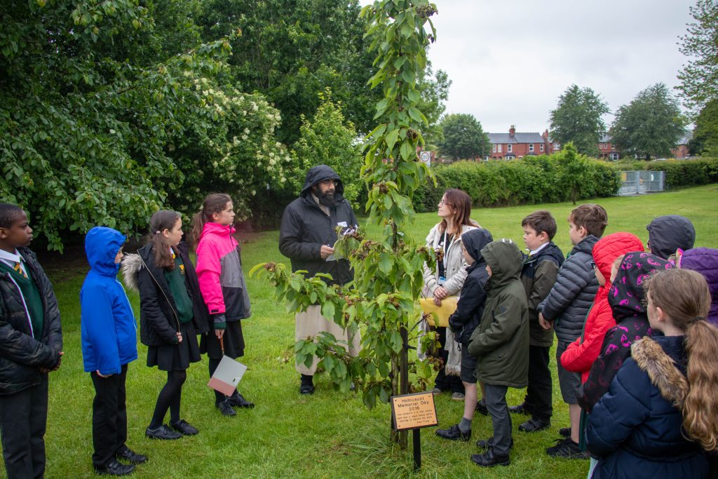 An image of Imam Sohayb Peerbhai with children from Woodside Primary School in Oswestry gathered around a cherry tree commemorating Ann Frank and the remembering the victims of the Holocaust. 